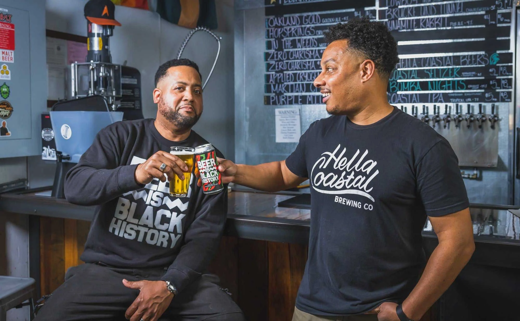 Two men smiling and toasting beer glasses in a brewery—perfect for branding photography with a chalkboard menu behind them.