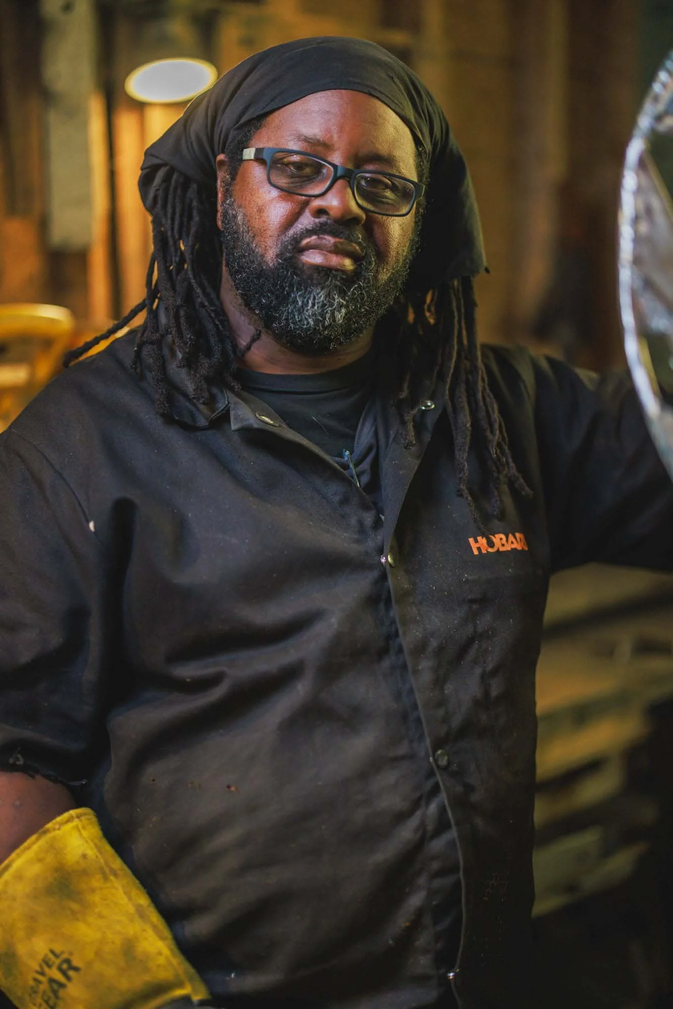 A man with glasses and dreadlocks stands indoors, holding a yellow work glove—an authentic moment