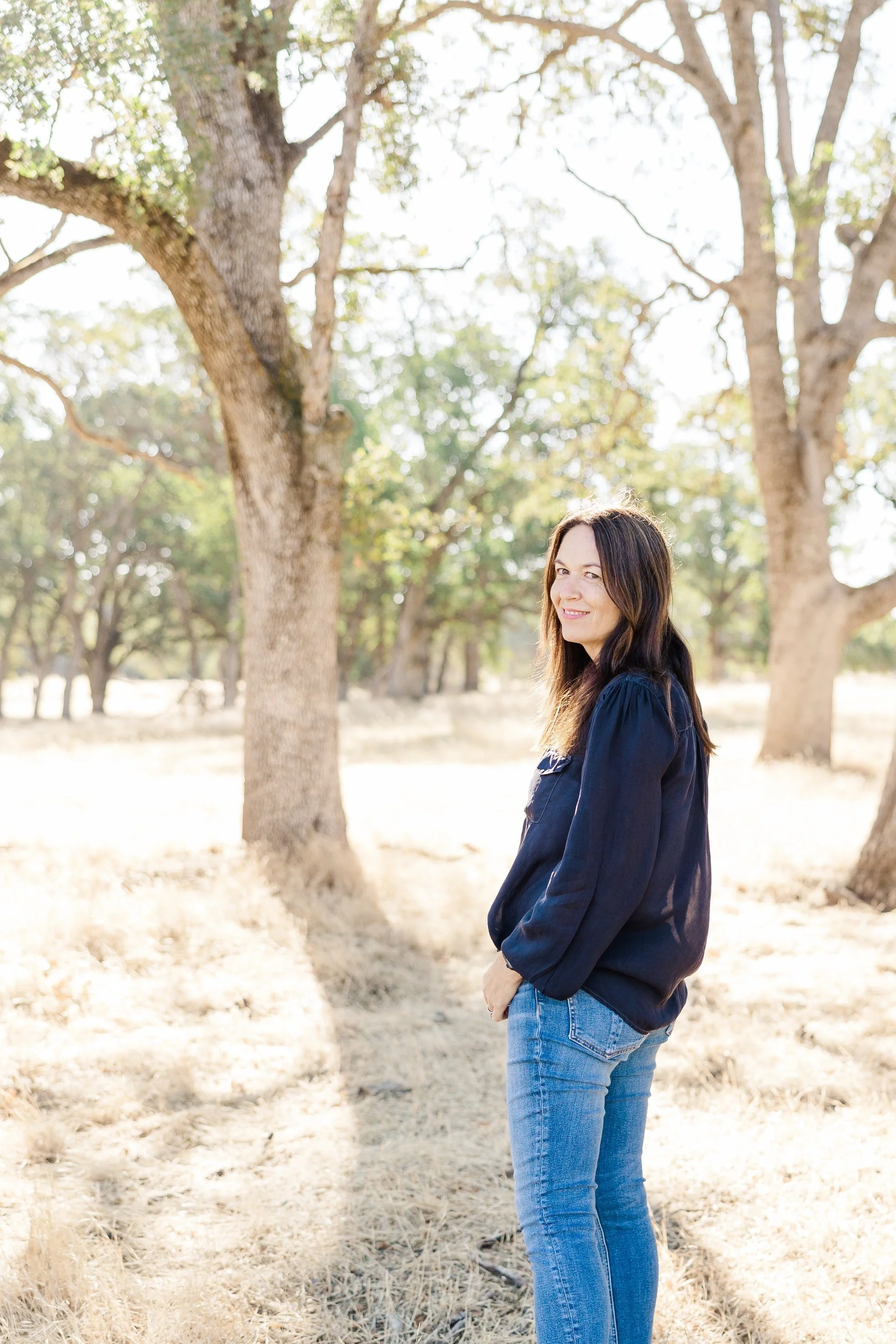 A woman with long dark hair wearing a navy jacket and blue jeans standing in a sunlit park with trees and dry grass, smiling at the camera.