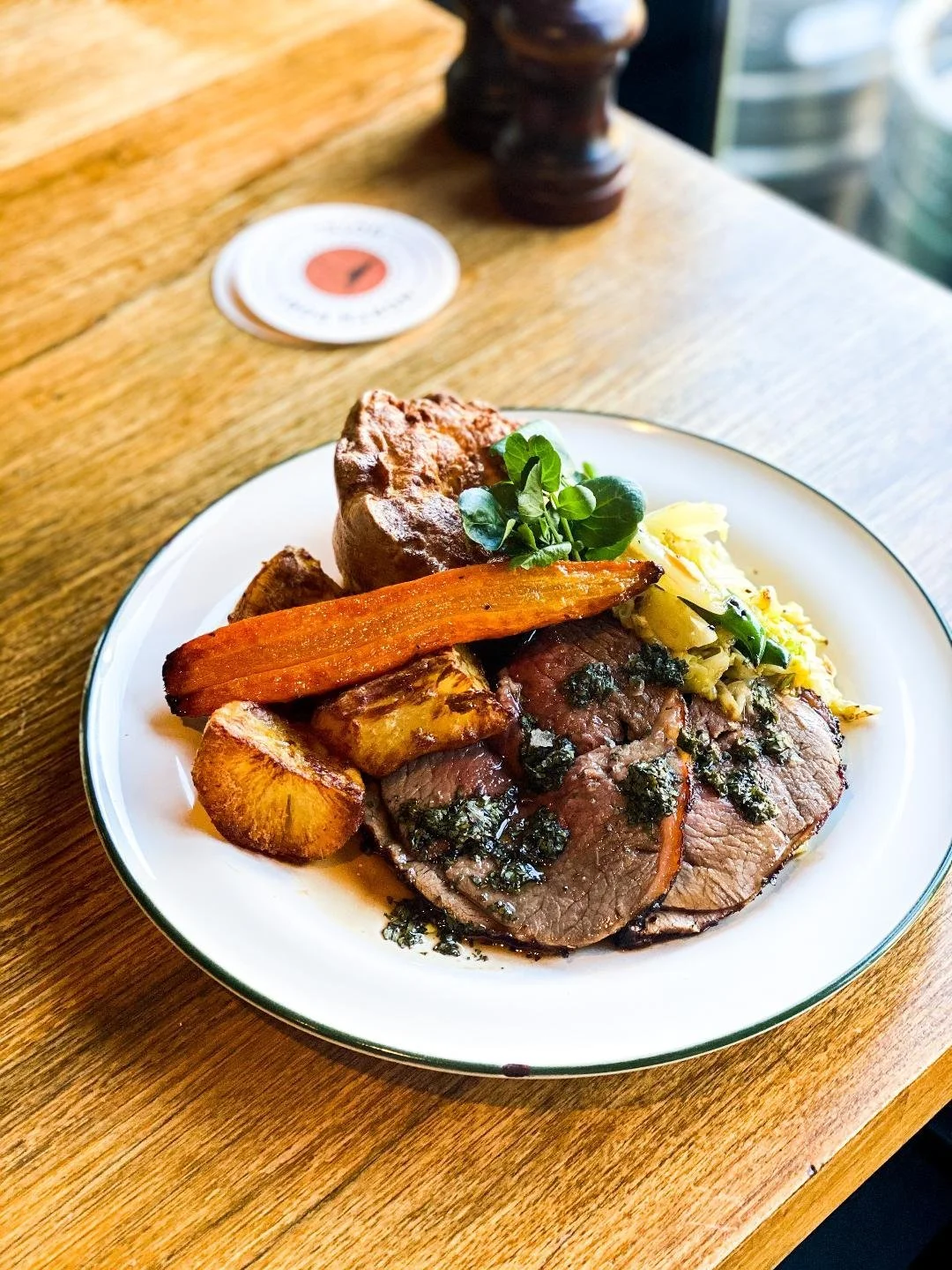 Plate with roasted beef slices topped with herbs, roasted vegetables including carrots, potatoes, and parsnips, a serving of mashed potatoes with greens, and a piece of bread, on a wooden table.