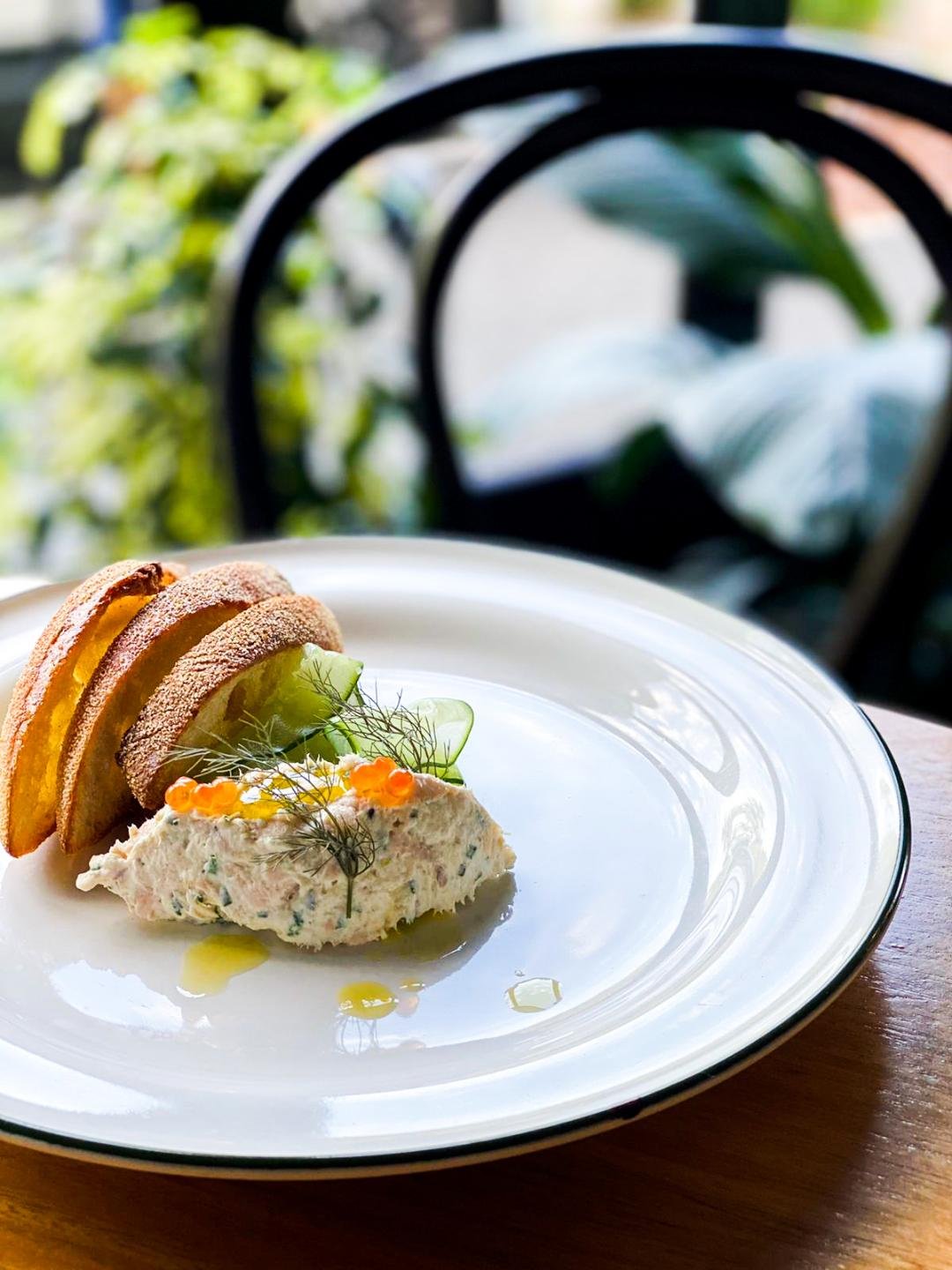 A white plate with a gourmet appetizer featuring two small sandwiches, a dollop of spread garnished with dill and orange roe, sliced cucumber, and pickles, drizzled with olive oil, placed on a wooden table with a blurred outdoor background.