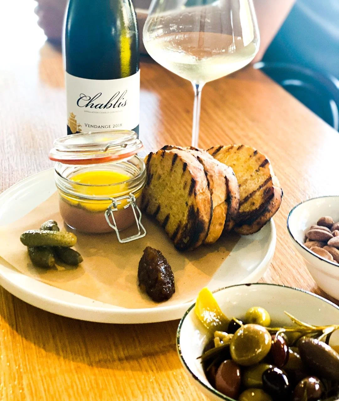 A plate with slices of grilled bread, a small jar of butter, pickles, and a dark chutney on a wooden table. A bottle of Chablis white wine and a glass of wine are in the background. There are small bowls with olives and nuts nearby.