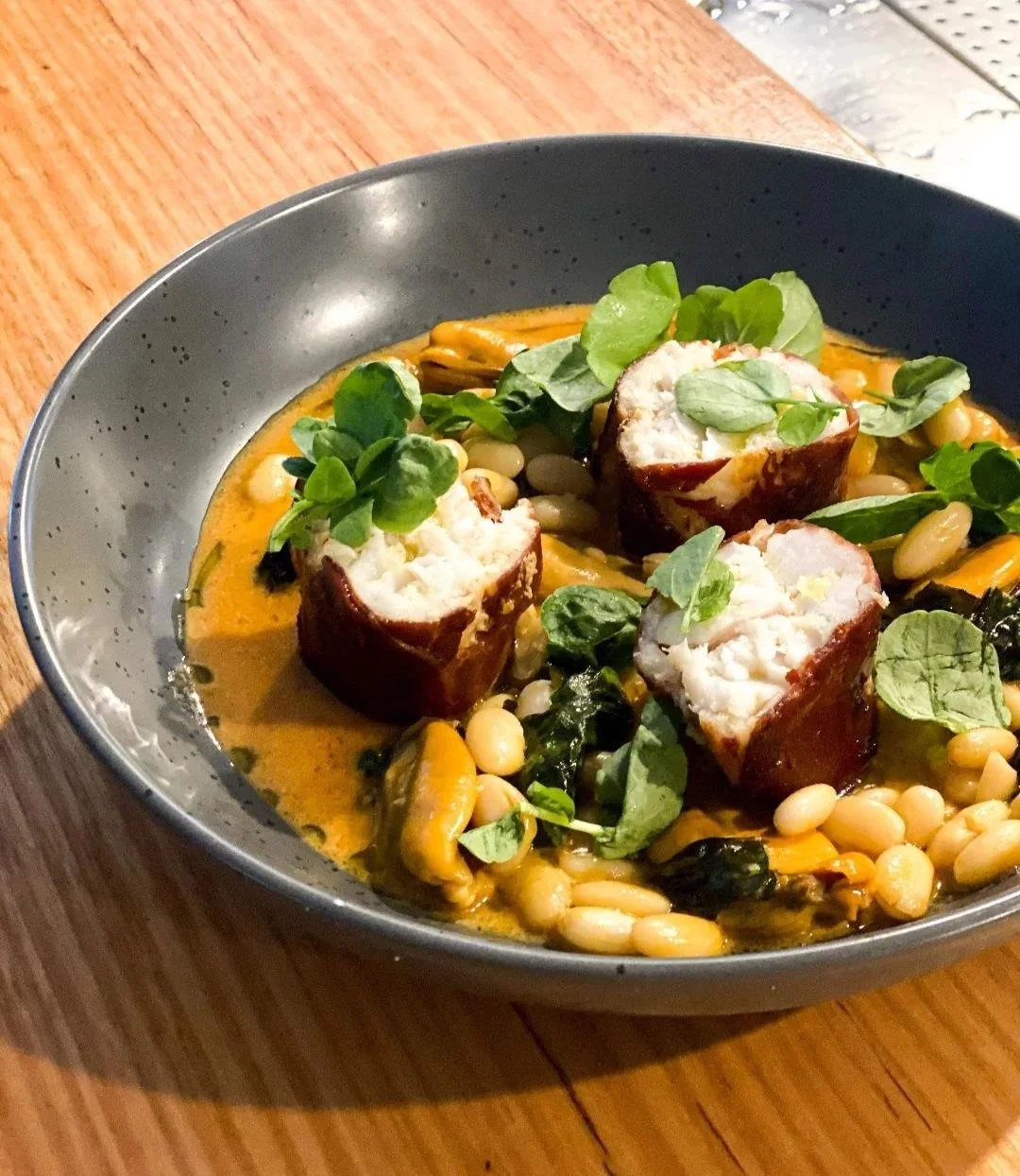 Creamy soup or curry with three bacon-wrapped stuffed chicken breasts, garnished with fresh herbs, served in a black bowl on a wooden table.