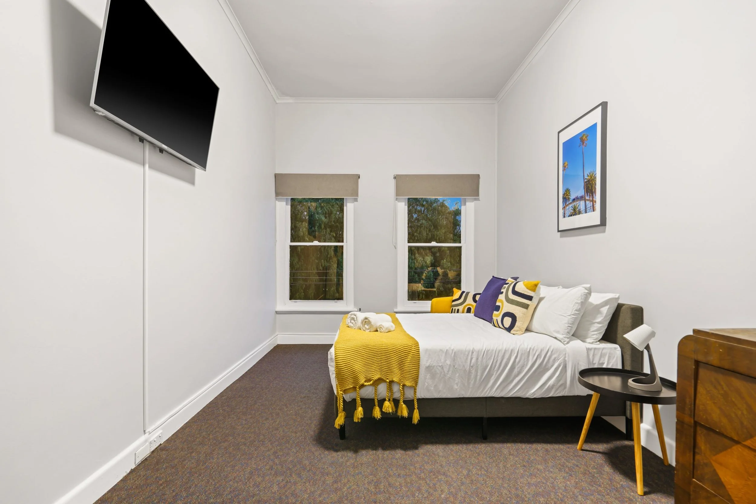 A bedroom with two windows, a bed with colorful pillows and a yellow blanket, a small round black side table with a lamp, and a framed picture of palm trees on the white wall.