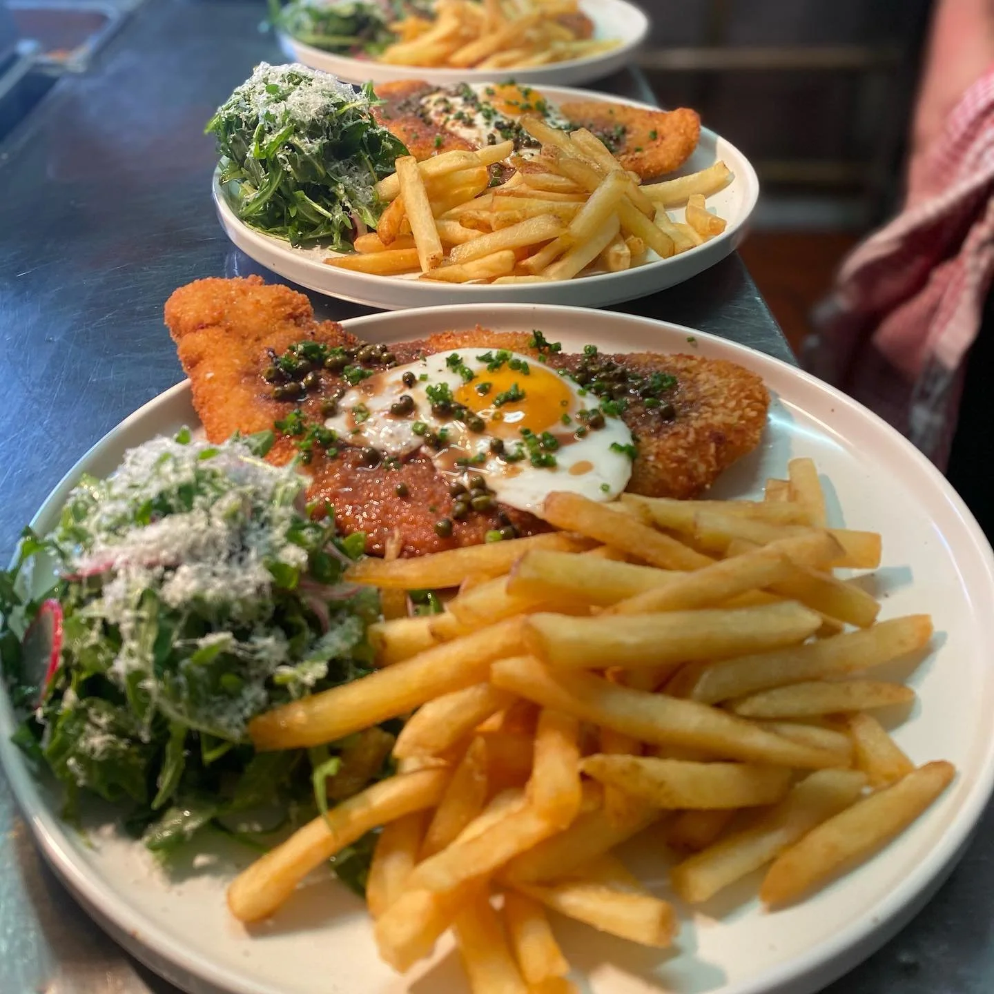 Plates with French fries, salad with shredded cheese, fried fish topped with a sunny-side-up egg, and green herbs.