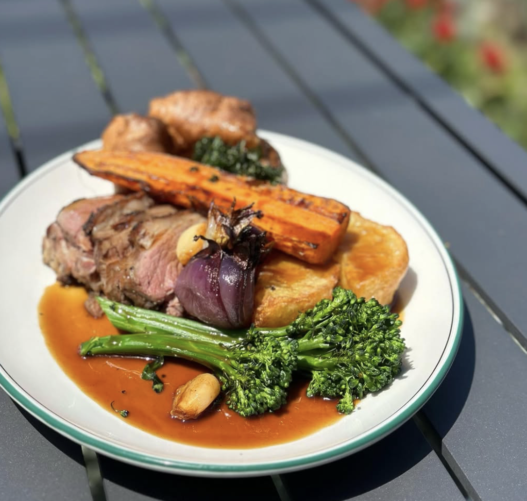 Plate of cooked meat, roasted carrots, broccoli, garlic, and roasted potatoes with gravy on a black outdoor table.
