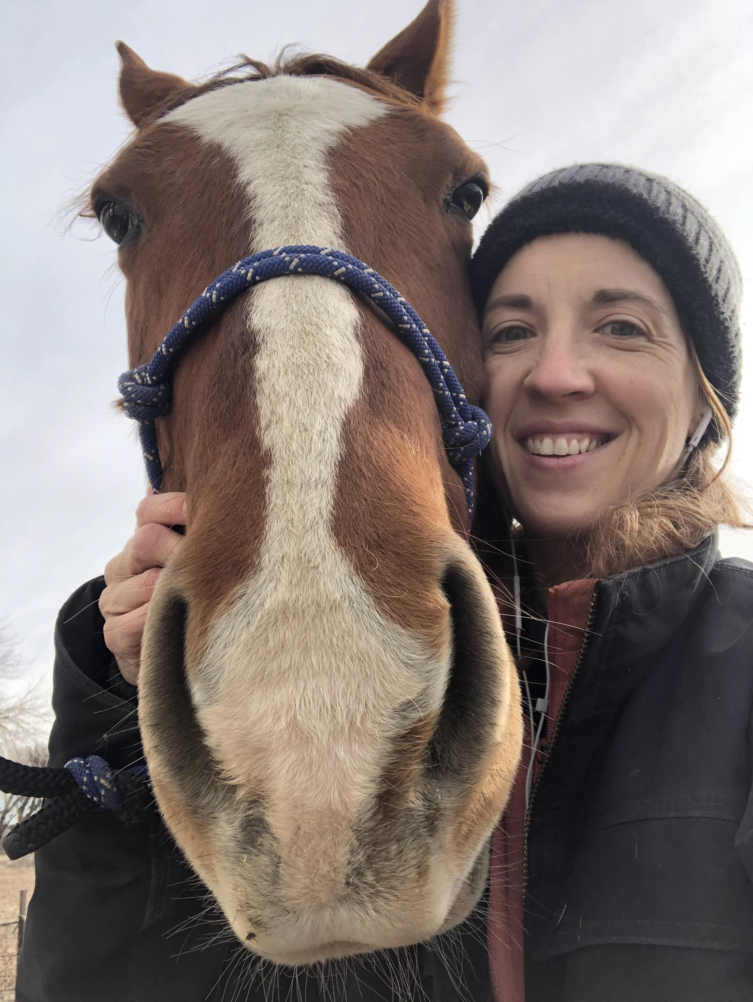 Woman standing with horse, horse's face covering most of frame, woman touching horses neck and smiling, horse reddish-brown with large white stripe up front of his face and white muzzle
