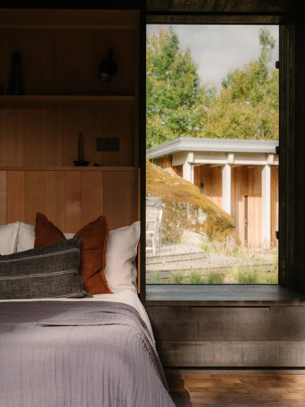 A bedroom with a bed, pillows, and a large window showing an outdoor scene with trees and a building.