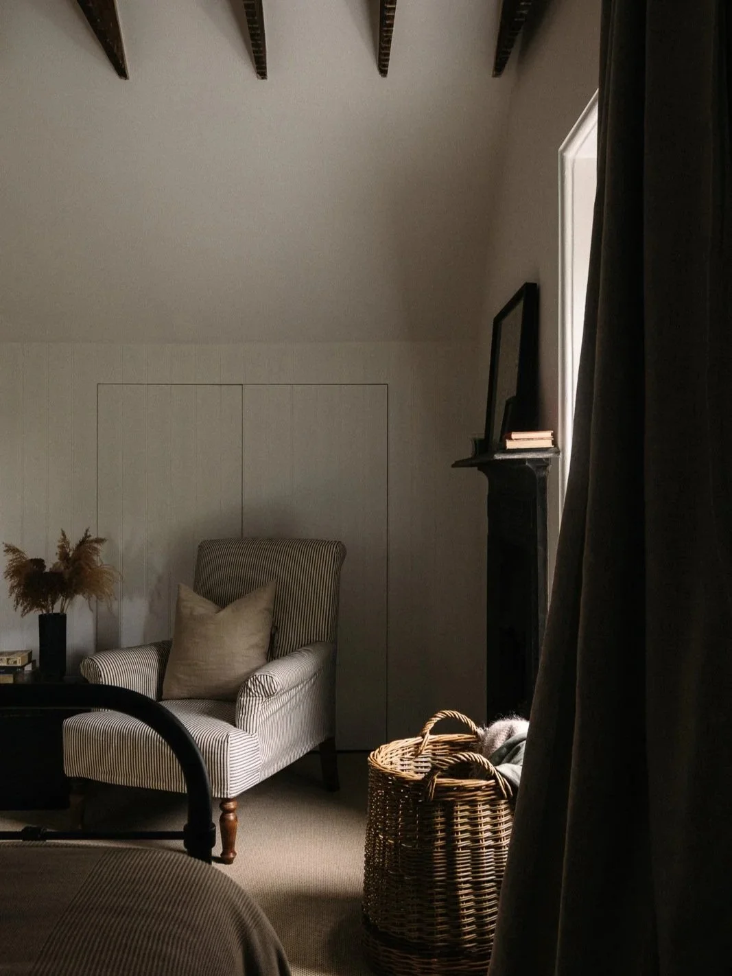 A cozy living room with a striped armchair, a beige pillow, a dark side table with dried flowers, a woven basket, a window with dark curtains, and a fireplace with a mirror and books on top.