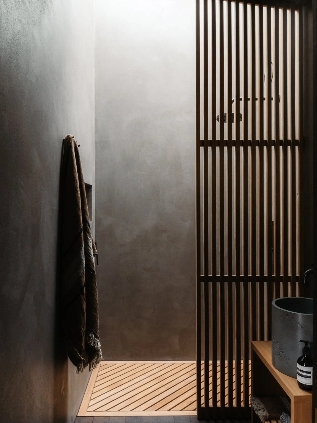 Minimalist bathroom with a walk-in shower, wooden slat partition, a towel hanging on the wall, and a concrete sink on a wooden shelf.