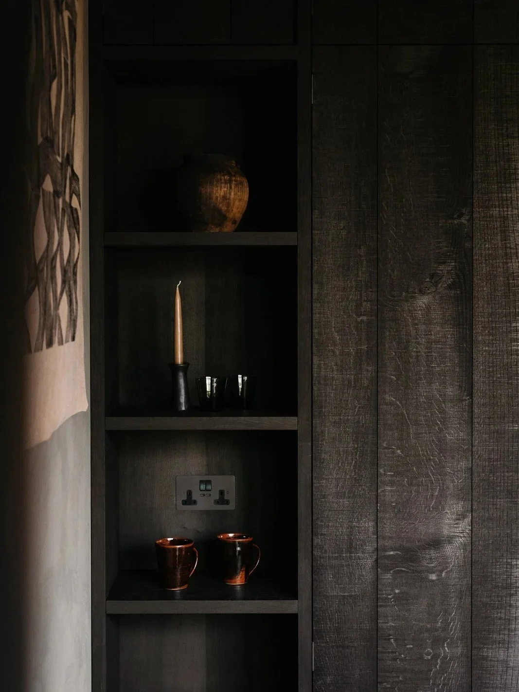 Dark wooden shelf with decorative items, including a vase, candle, black bowls, electrical outlets, and two small ceramic cups, beside a textured wall.