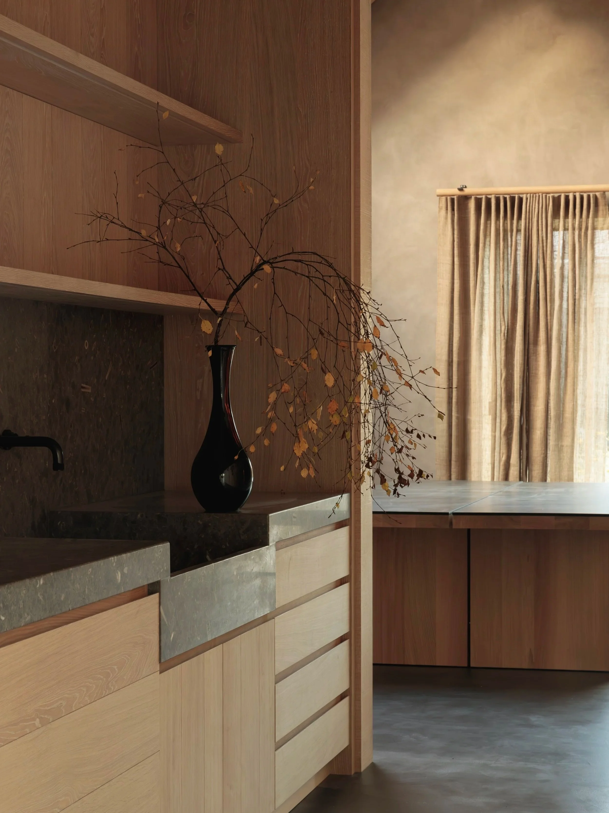 A minimalist kitchen with natural wood cabinets, a dark vase with dried branches and leaves, beige curtains, and a window with daylight