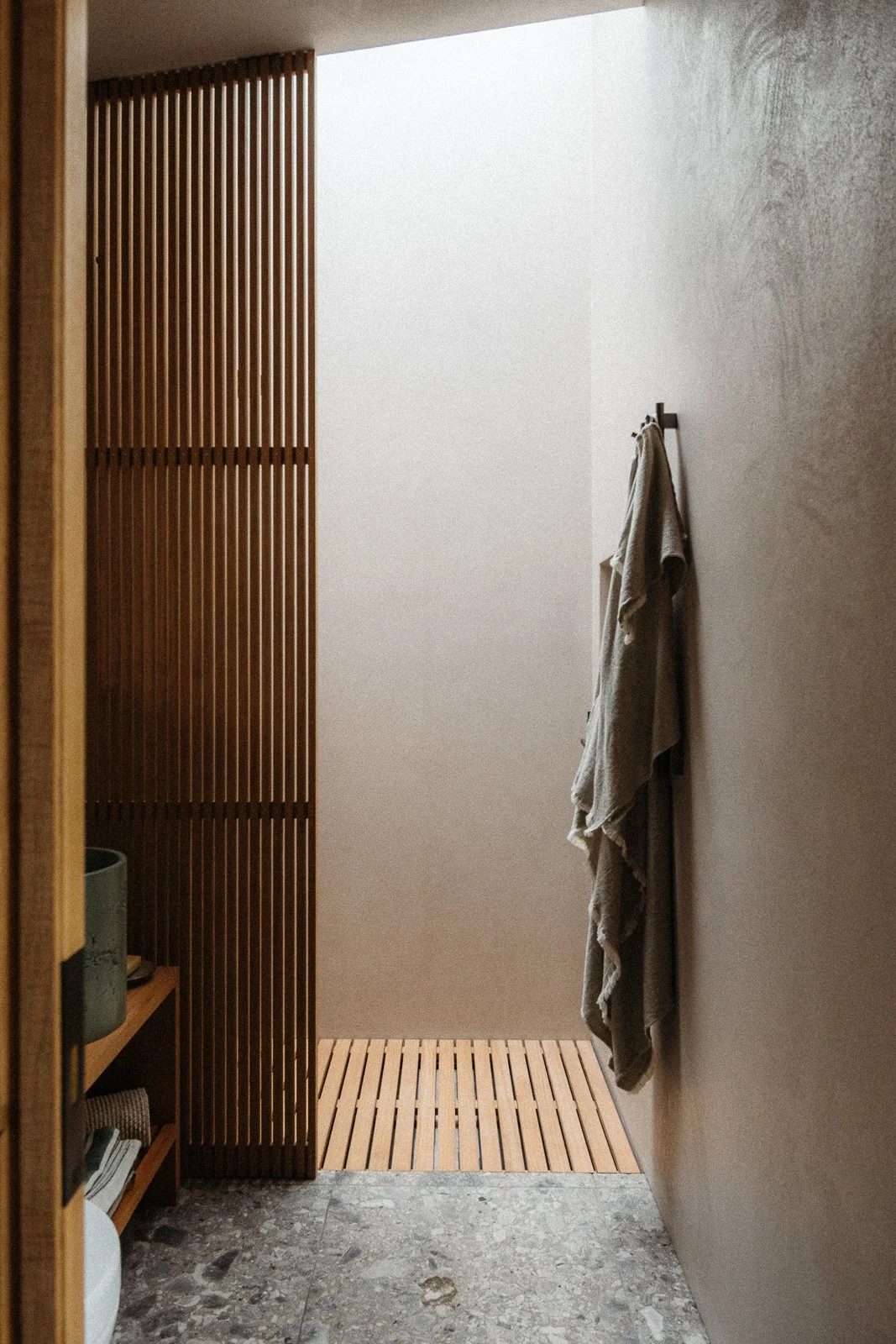 A minimalist bathroom with a wooden slatted partition, a towel hanging on an wall hook, and a small wooden mat on a stone floor.