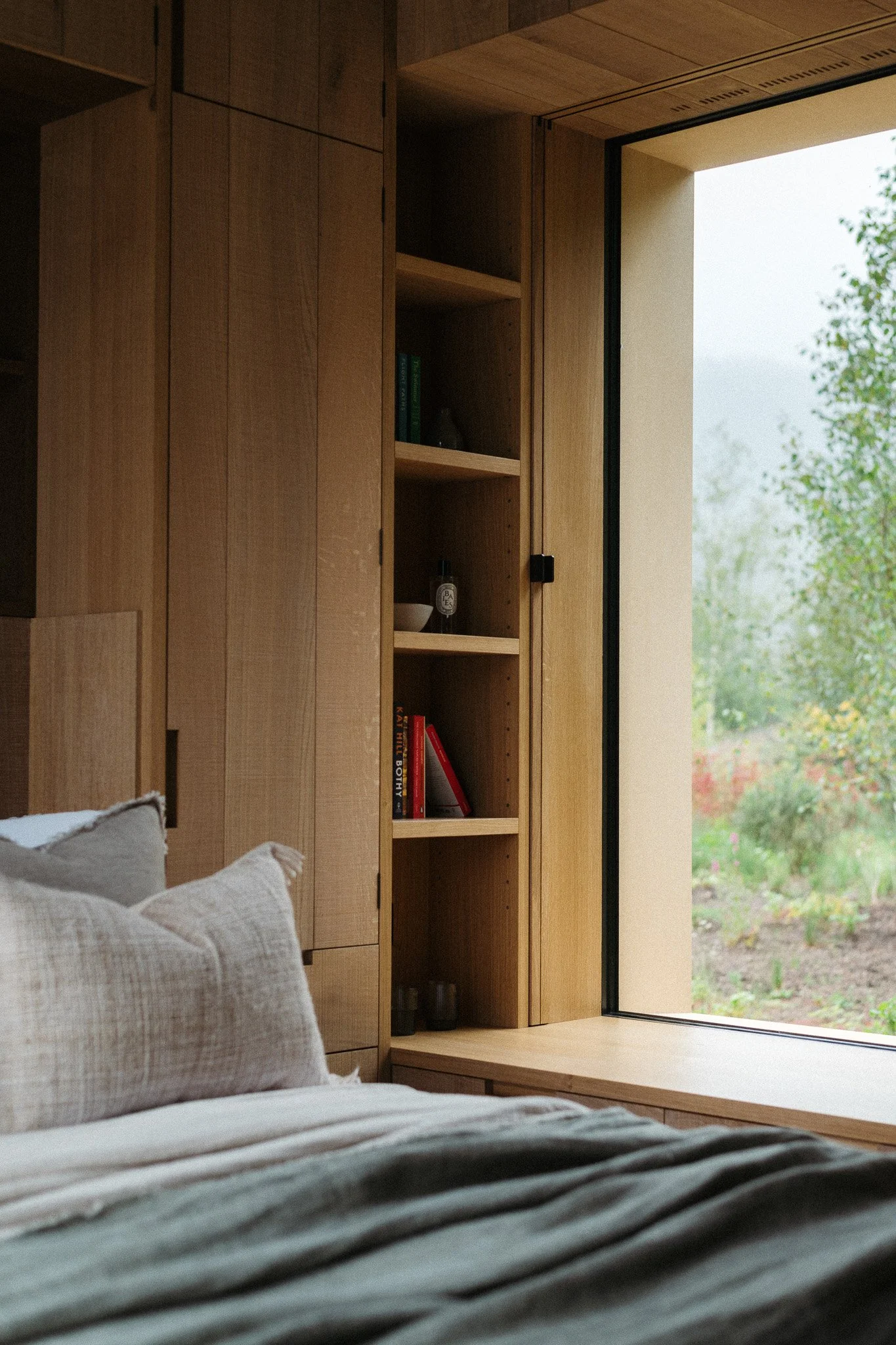 A bedroom with a bed in the foreground and a wooden bookshelf with a window view in the background.