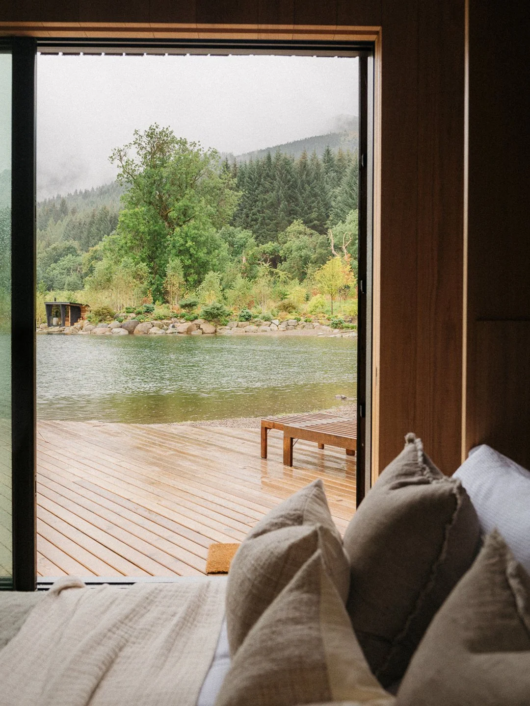 View of a lake with a wooden deck and bench, surrounded by trees and mountains, seen from inside a room with beige pillows