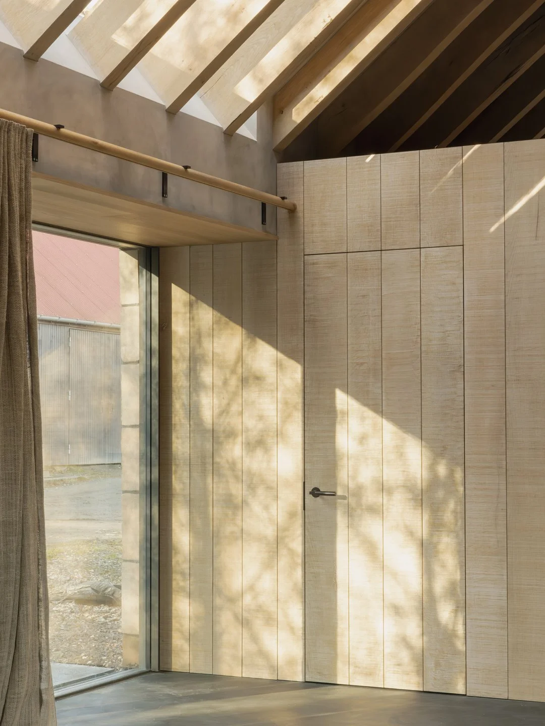 Interior of a room with light-colored wooden walls, large window with sunlight casting shadows of tree branches, and a door with a black handle. Part of a curtain and a wooden railing are visible.