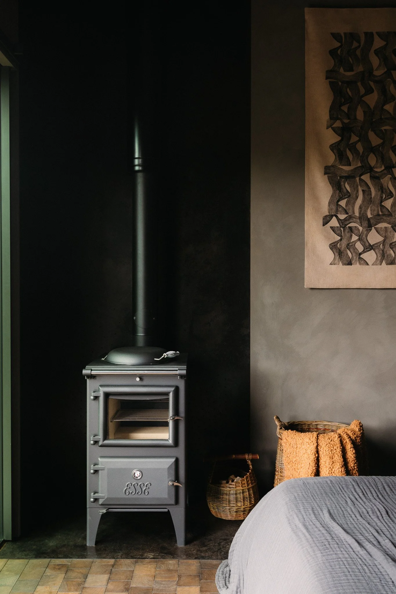 A gray wood-burning stove with a black pipe, a wicker basket, and an orange towel in a room with dark walls and modern artwork.