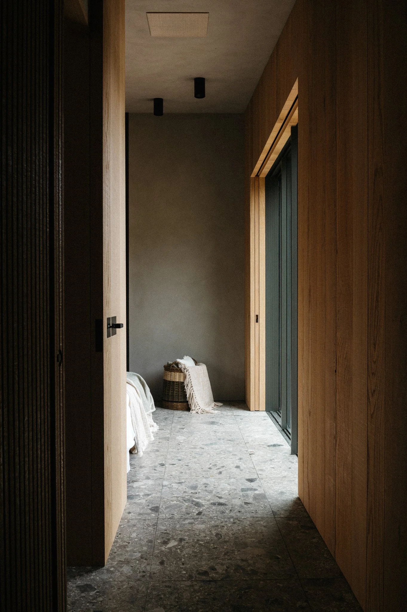 A minimalist hallway with wooden walls, gray tiled flooring, and a gray back wall. There are two black ceiling lights, a basket with white towels, and a woven blanket on a bench on the left side.