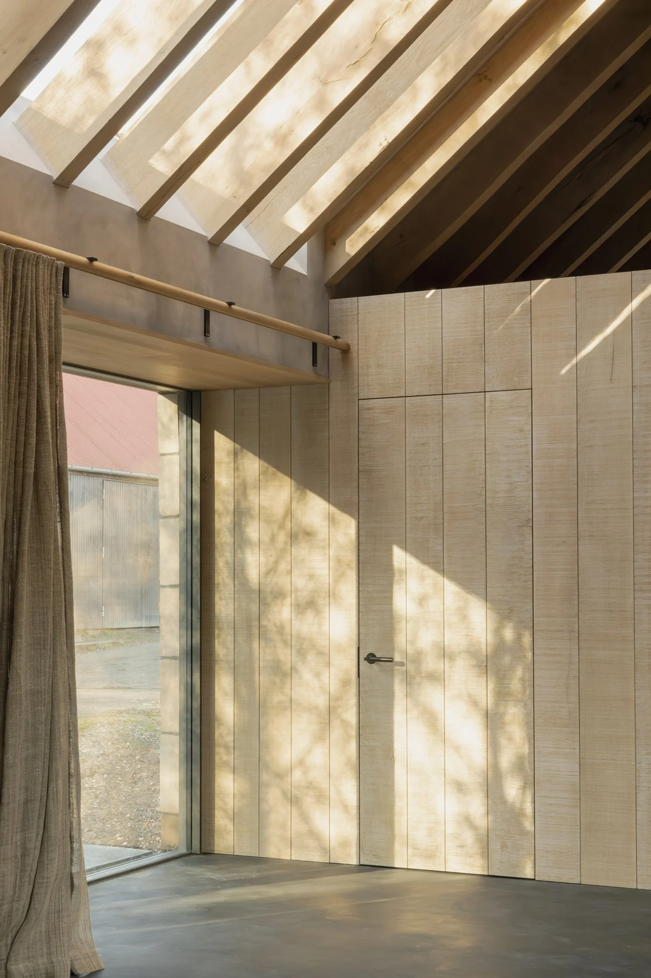 Interior view of a room with large glass sliding door, beige wall paneling, wooden beams in the ceiling, and sunlight casting shadows inside.
