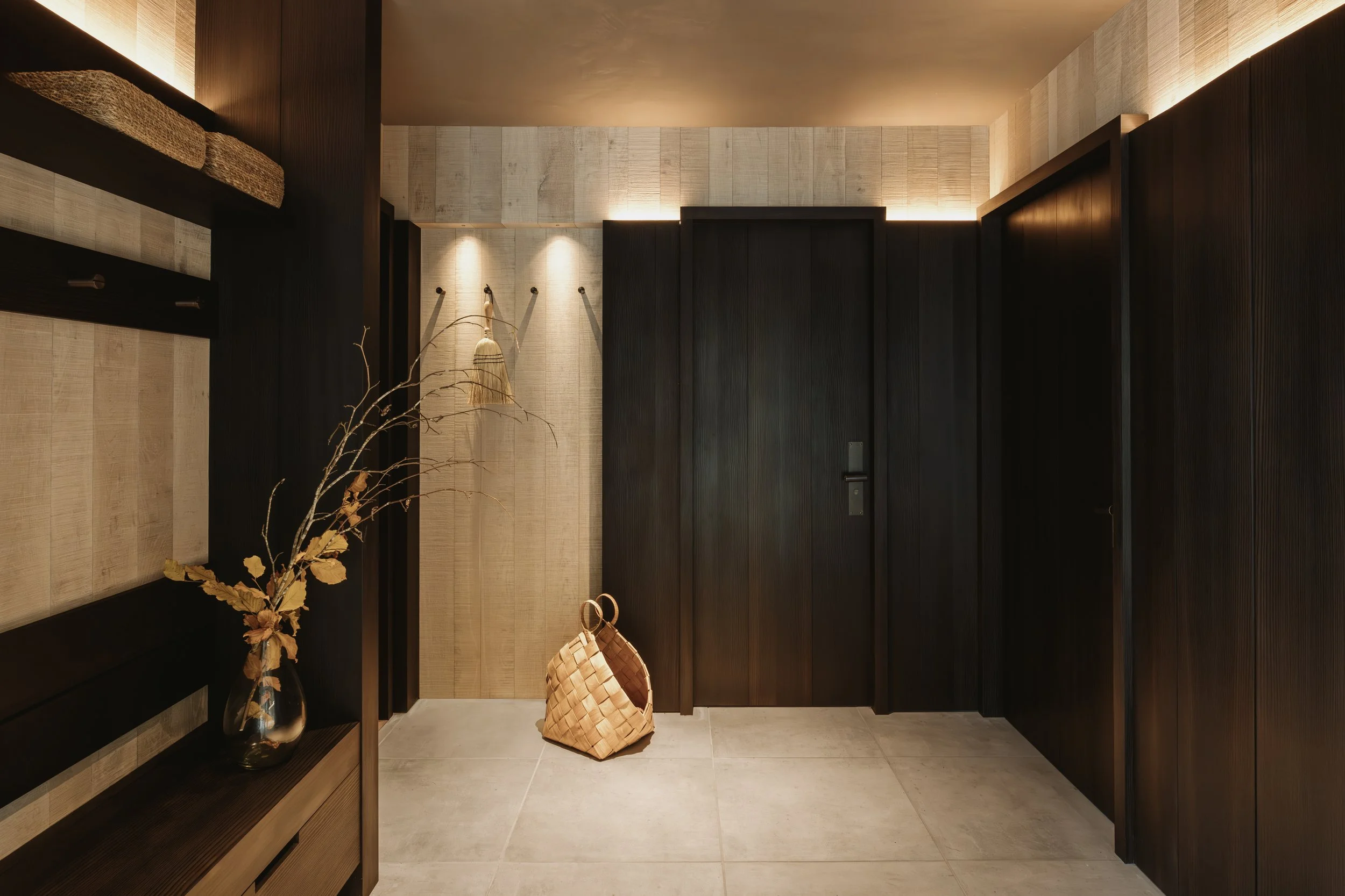 Modern entryway with dark wooden walls, beige tiles, a decorative branch in a glass vase, a woven basket, and wall hooks with a small hanging bag.