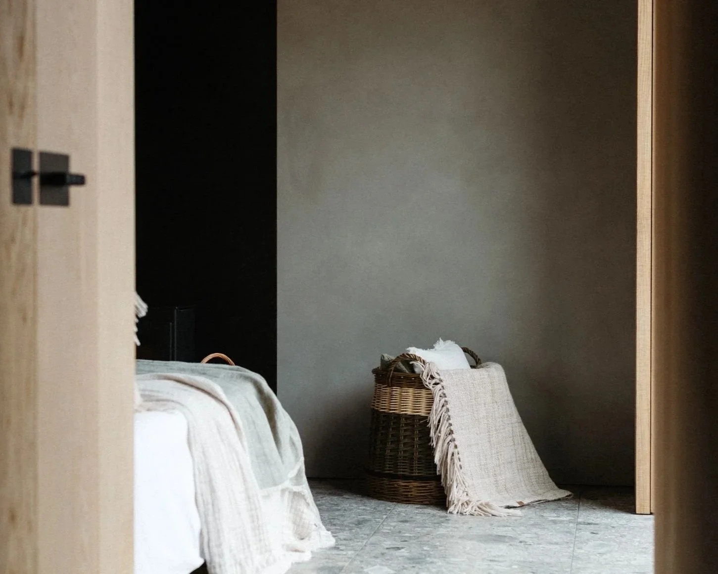 A cozy bedroom corner with a bed covered in neutral-toned blankets and pillows, a wicker basket with a cloth draped over it, and a textured gray wall in the background.