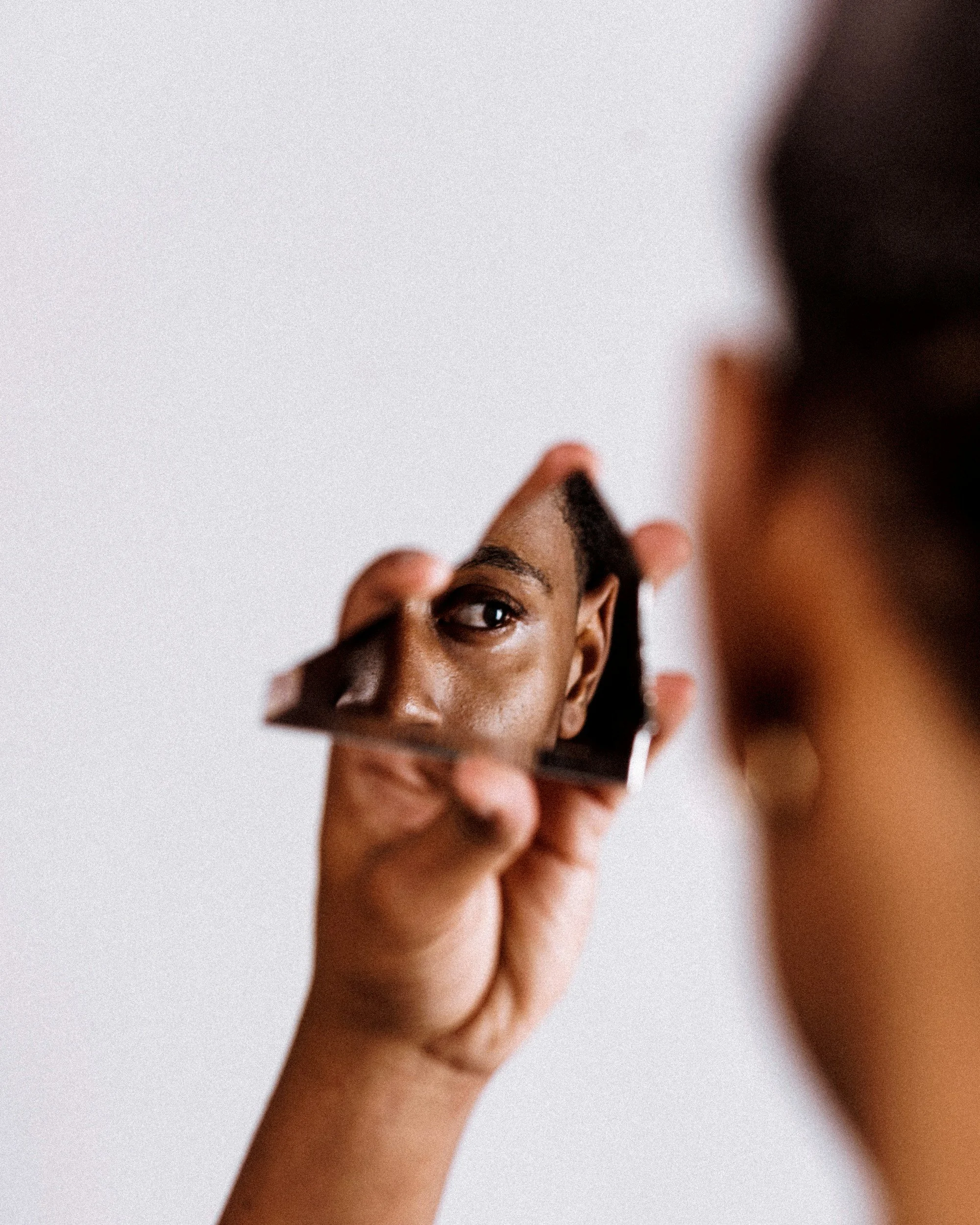 Close-up of a person holding a mirror reflecting their eye and part of their face, with another person's head partially visible in the background.