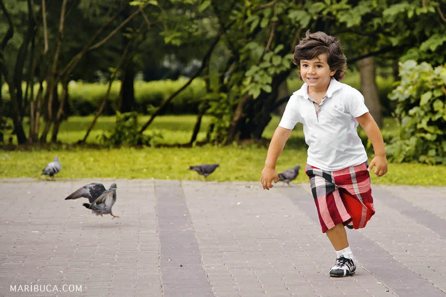 Happy 5 year old boy running in the park on a background of pigeons and green trees.