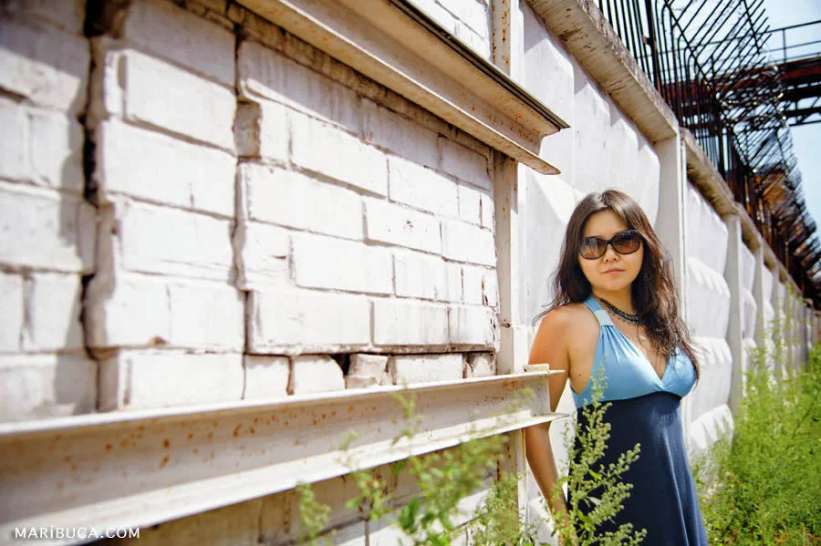 The girl in the blue dress and sunglasses standing against a white fence.