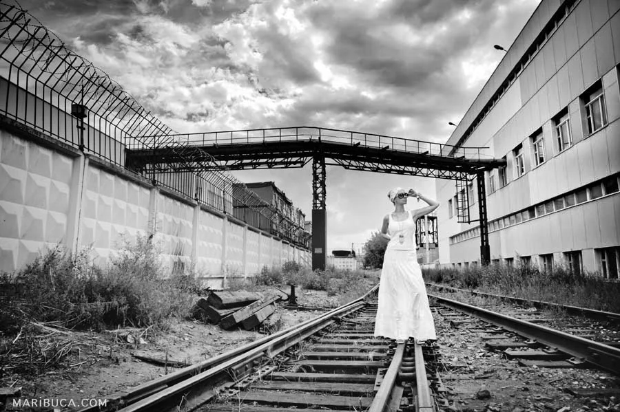 A black and white photo of an Industrial view of a factory with old rails and a girl standing in a white T-shirt and white long skirt at Campbell.