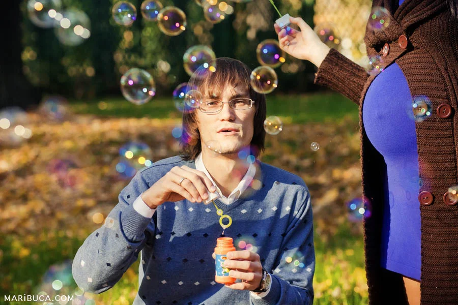 The guy in a light blue sweater launches colored soap bubbles and a pregnant wife standing to his left.