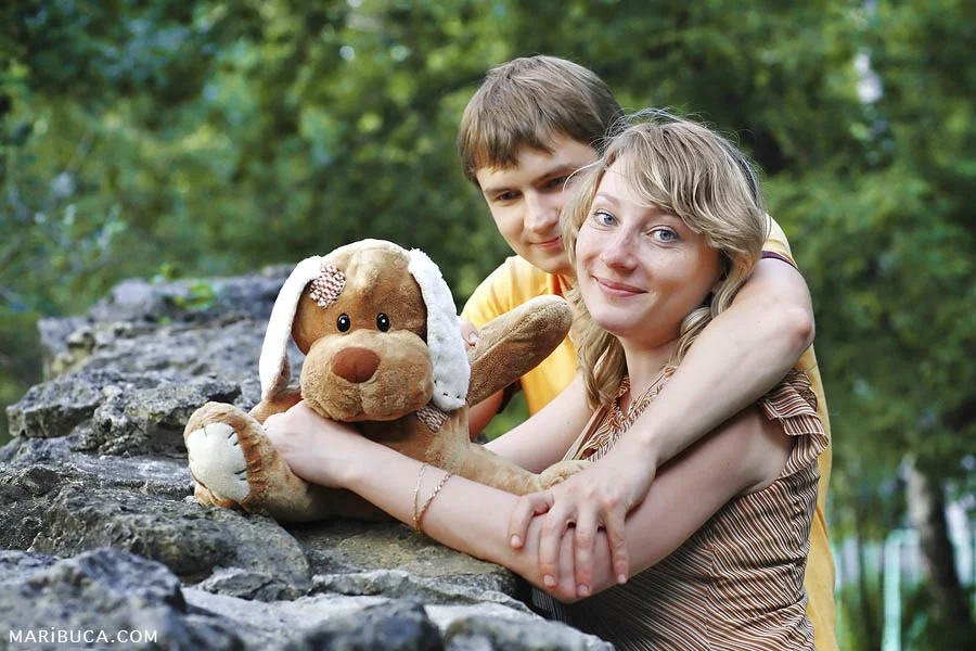 Photo session in Palo Alto where a couple hugs each other and a soft brown-white toy dog against the background of green trees.