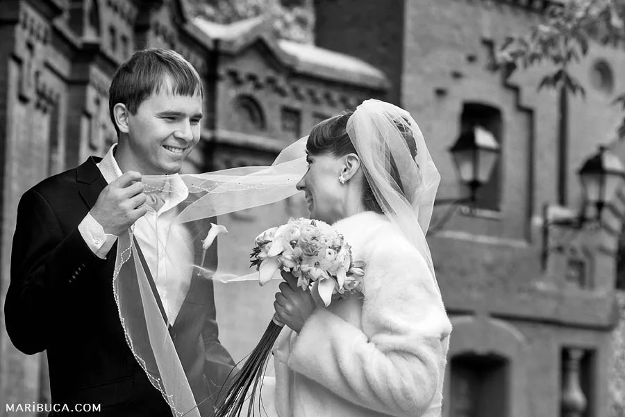 The groom looks tenderly at the bride and holds her veil against the background of the church.