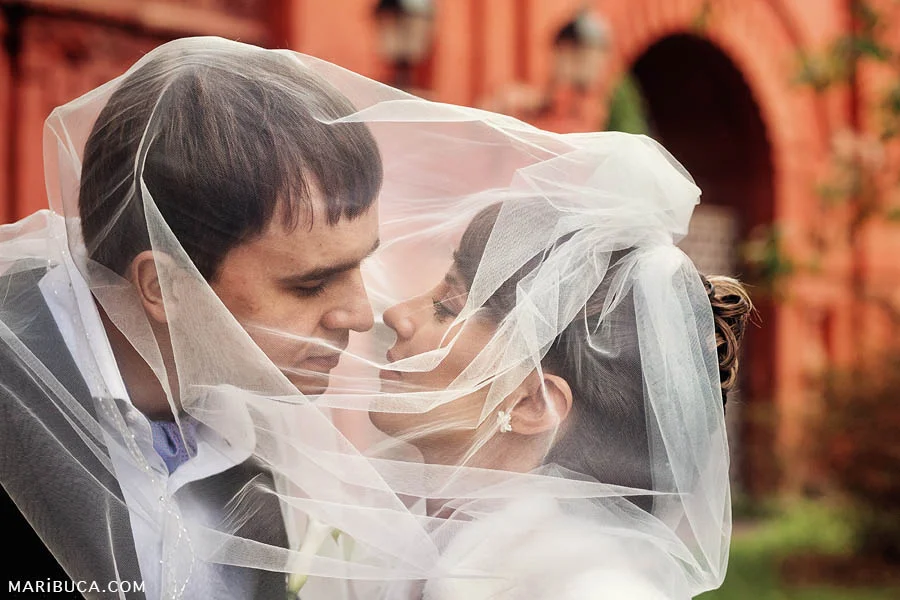 The bride and groom almost kissed and both are covered by a veil of the bride on a background of a red wall.