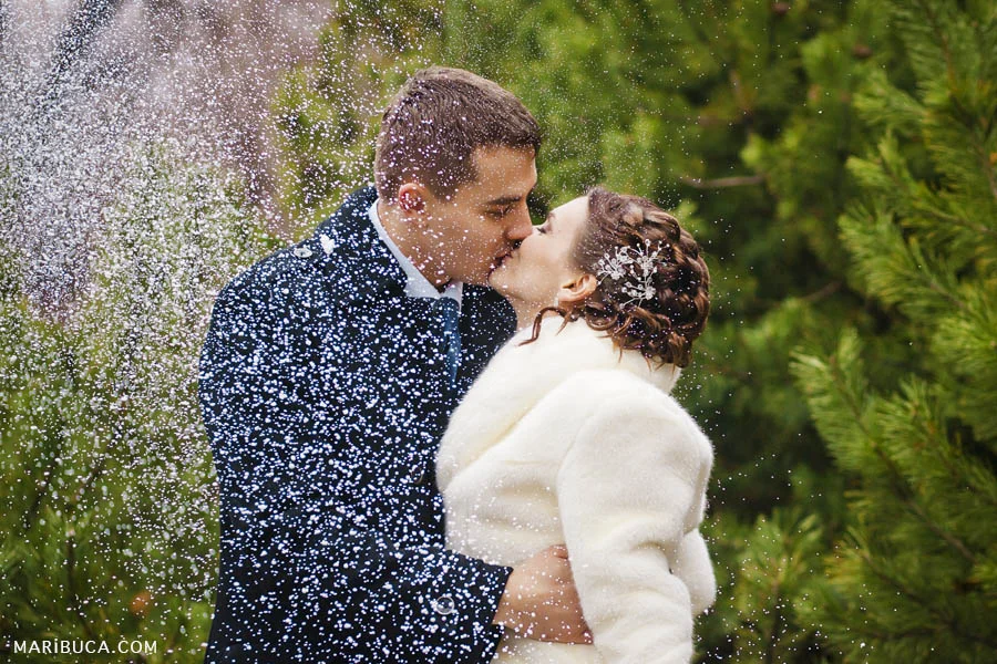 the bride and groom kiss each other and it snows in winter weather on the background of the trees.