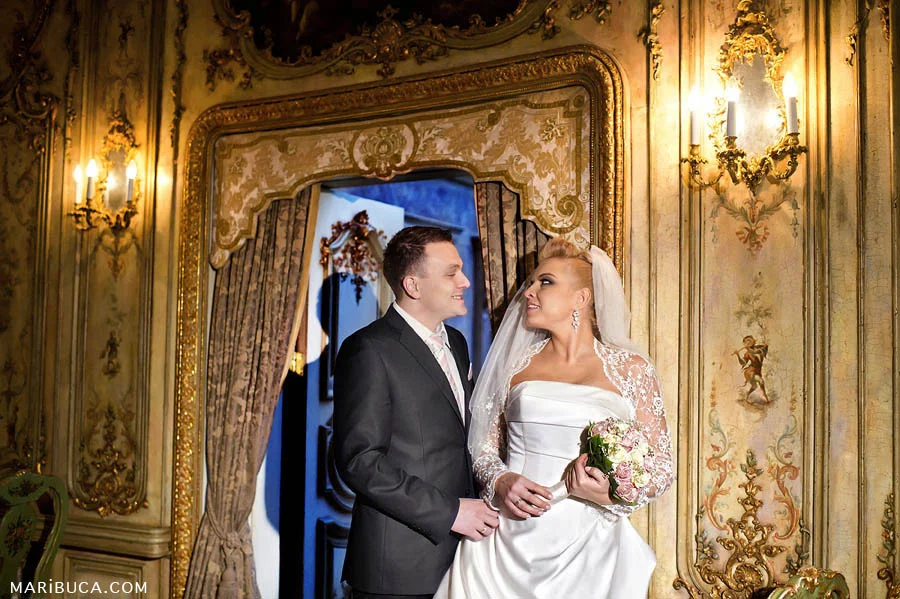 The groom and bride hold and look each other in front of golden beautiful wall with candelabra