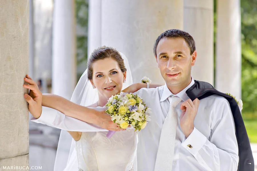 the groom holds a jacket over his shoulder, the bride leaned on the groom's hand and holds a light yellow bouquet against the white columns.