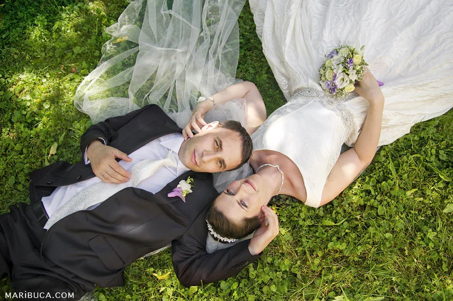 the bride and groom lie on their backs on the green grass and enjoy each other's company