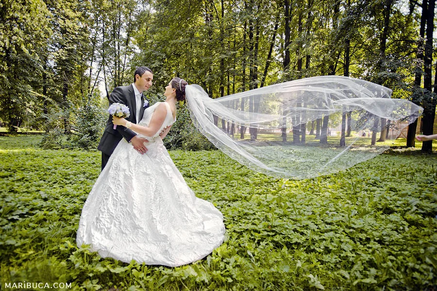 the groom to the bride looks like she flies a long white veil in the wind on a background of green grass and trees.