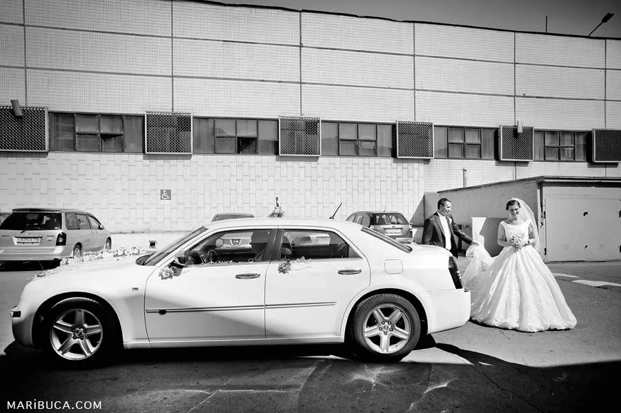 A black and white photograph of the bride and groom emerging from a white Bentley car against the backdrop of an industrial building.