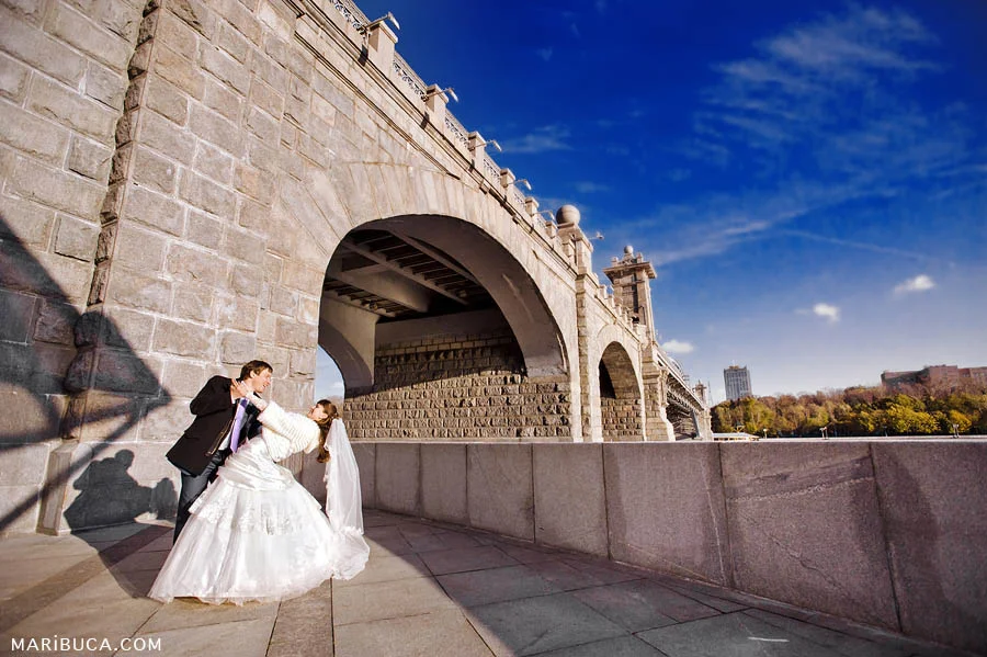The bride and groom are dancing against the backdrop of a huge bridge and a deep blue sky.