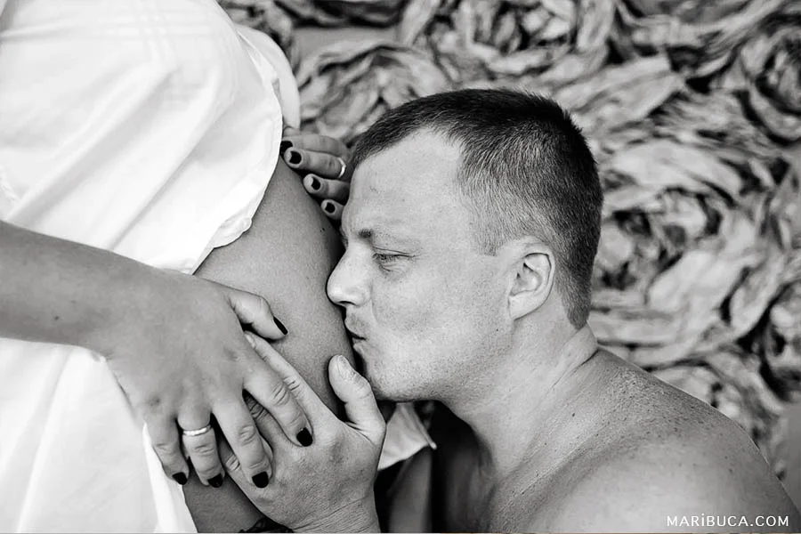 Black and white image of the future dad who is kissing wife’s belly and they are expecting the baby.