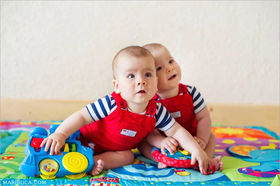eight-month-old boys twins in red jumpsuits sit each other surrounded by toys, colored soft rug and white wall