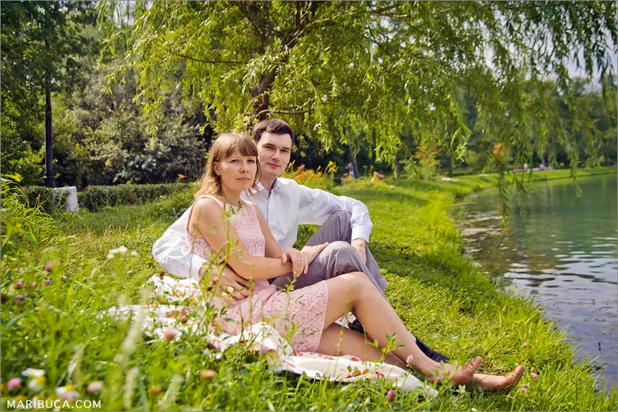 The fiance and fiancee are sitting in the green grass and pond around.