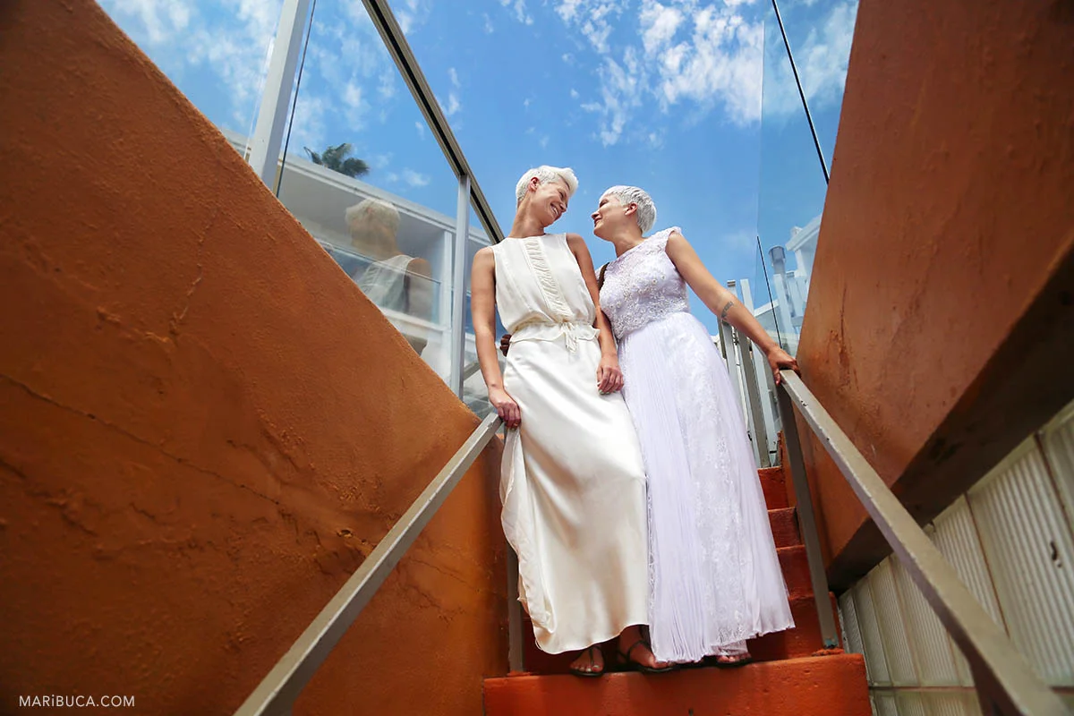 Two brides with white and ivory dress stay in the staircases and looking each other