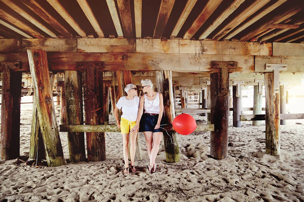 Two girls with sunglasses and the red balloon stay under the old wooden bridge speaking each other.