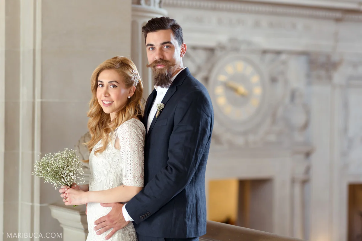 Just married couple stand in front of the clock on the wall in the 3rd floor City hall, San Francisco