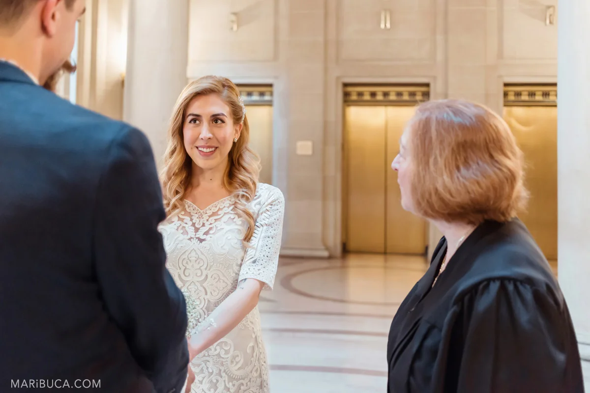The bride looks into the groom eyes and the officiant looks to the bride during the wedding ceremony in San Francisco city hall.