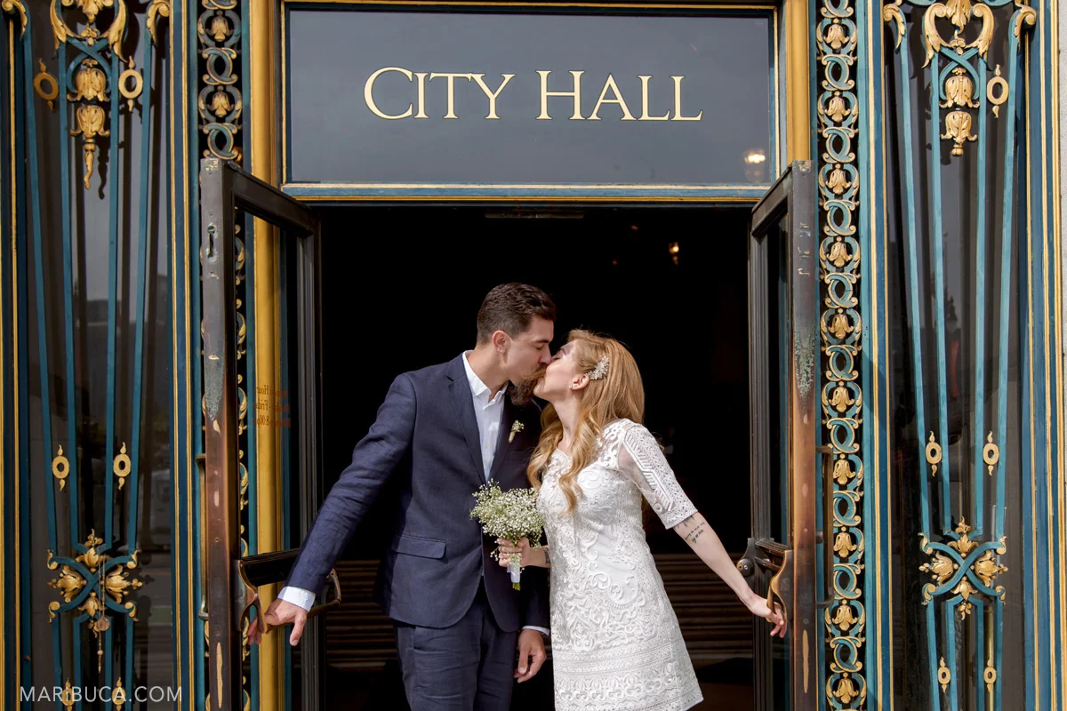 the groom in the blue suit and the bride in the white wedding dress hold the door handles and kiss leaving San Francisco City Hall. They just married!