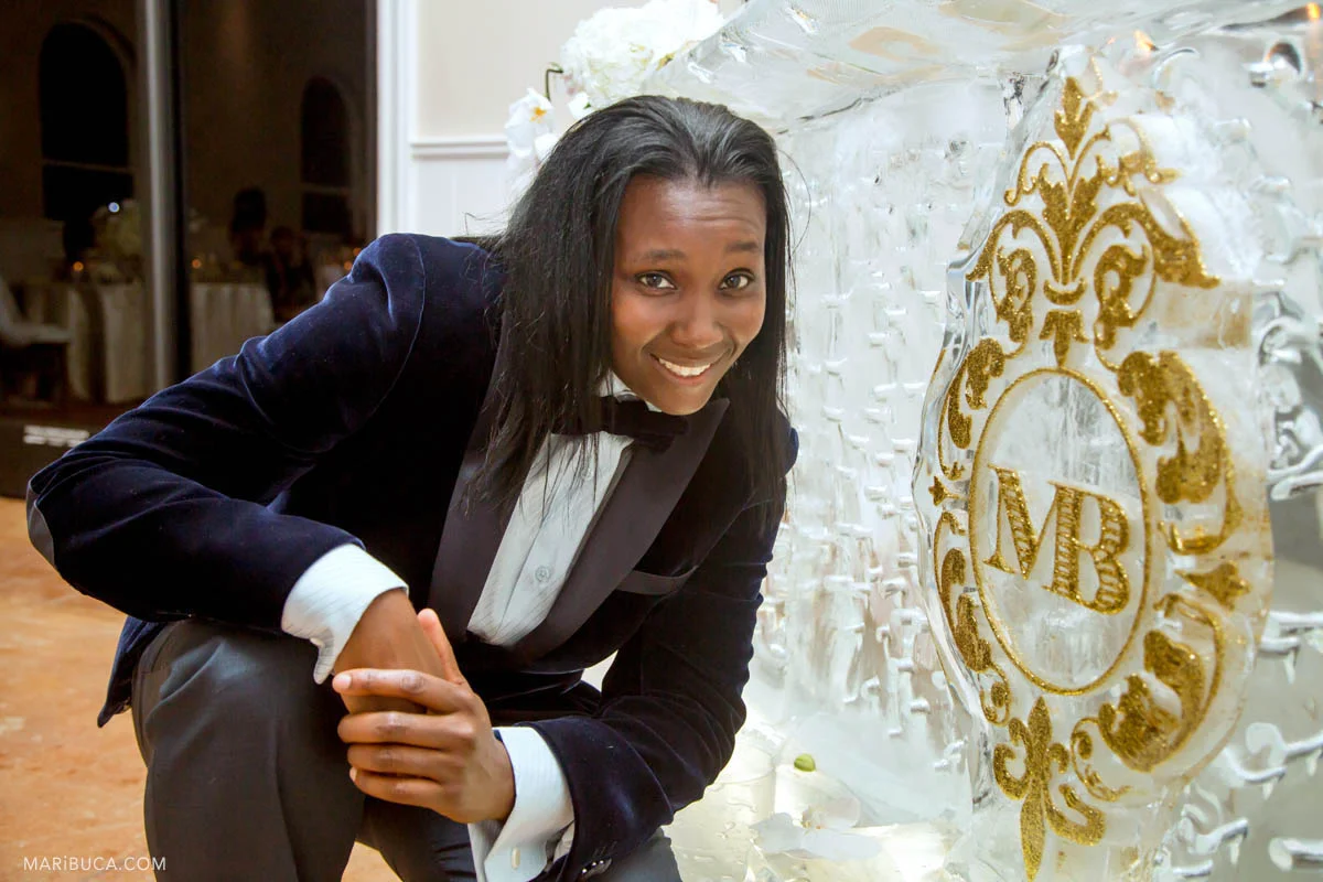 The groom girl in the dark velvet jacket smile next to the ice bar with golden sign MB