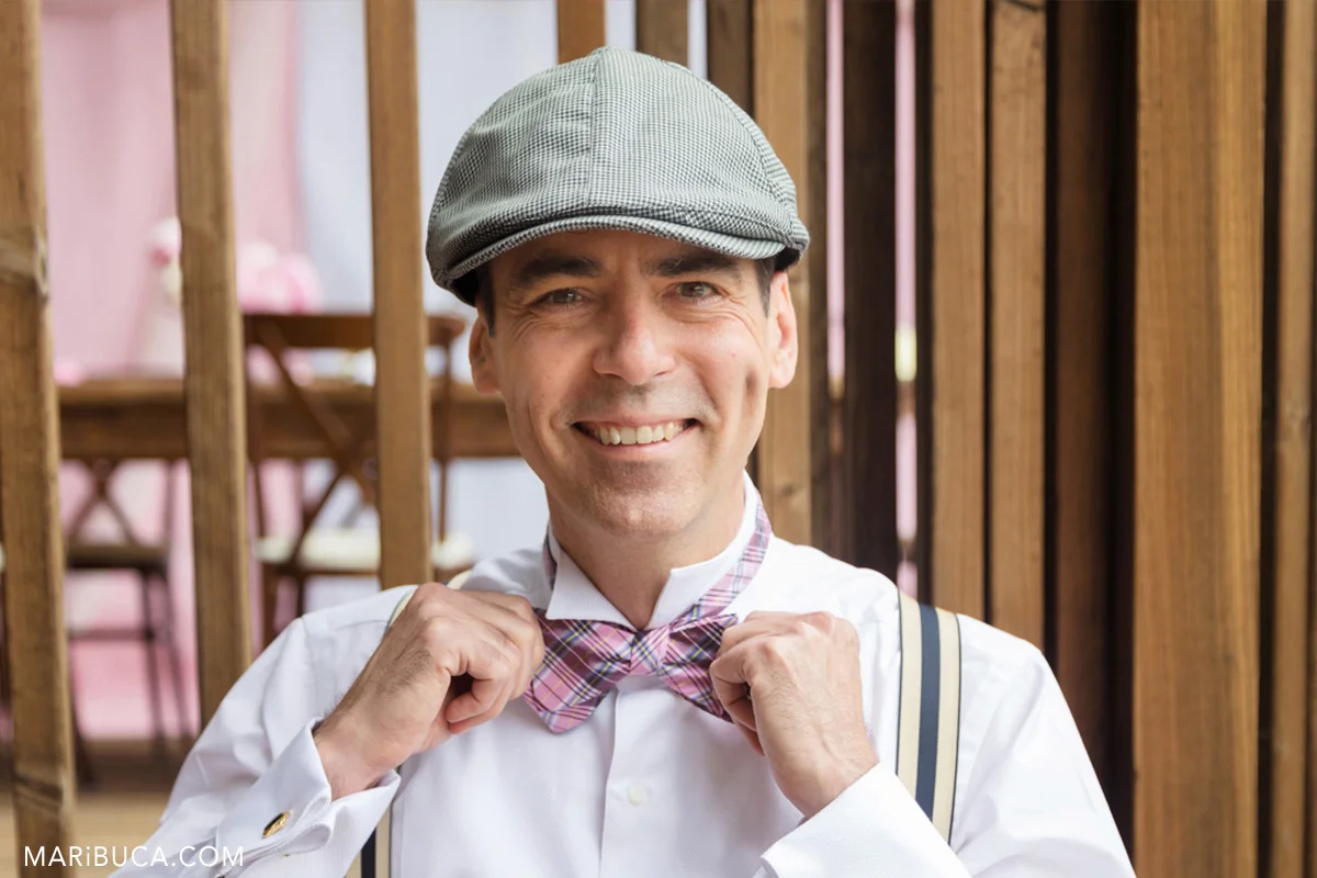 The groom smile and hold it his pink bow. He wears light black hat and white shirt in the getting ready.